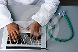 Close-up top view of unrecognizable African practitioner male doctor in white coat uniform working typing on laptop