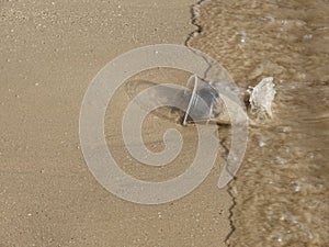 Close up top view of a Waste transparent plastic cup on the sand beach, a moment before rushed by sea wave, with copy space