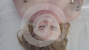 Close-up top view of young blond woman with grey eyes lying on couch with needles in face and sculp. Portrait of