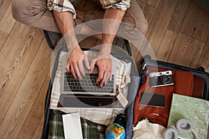 Close up of tourist, hands typing on laptop, opened suitcase with clothes for holiday, man packs luggage