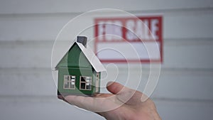 Close-up toy house in female Caucasian hand with blurred For sale announcement on garage door at background