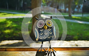 A close-up toy of a surprised blue owl with multi-colored eyes stands on a piece of wood in the light of the evening sun. The