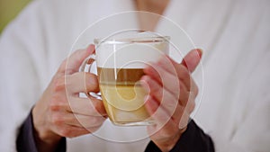 Close-up transparent coffee cup with cappuccino in mature female hands. Unrecognizable Caucasian woman in white bathrobe