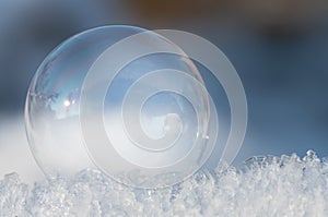 Close-up of a transparent and glowing soap bubble lying on glittering snow. The soap bubble looks like a glass ball. The