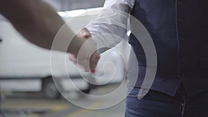 Close-up of two caucasian men shaking hands in car repair shop. Male hands at the background of vehicles. Car business
