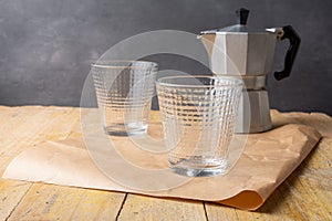 Close-up of two empty glasses with gray coffee pot on brown paper on wooden table, gray background, horizontal