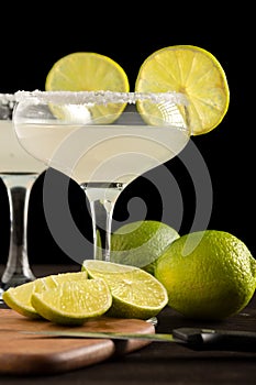 Close-up of two glasses with margarita cocktail, on dark wooden table with table, knife, limes and lemons, black background