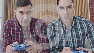 Close-up of two identical twin brothers looking at camera and using game consoles. Cheerful men playing video game on