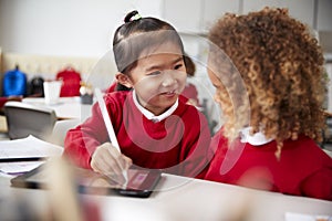 Close up of two kindergarten schoolgirls wearing school uniforms, sitting at a desk in a classroom using a tablet computer and sty