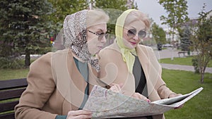 Close-up of two mature Caucasian women in headscarves and sunglasses sitting on the bench and examining the map. Senior