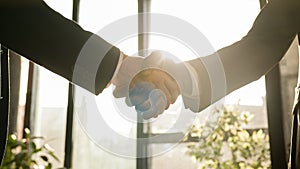 Close-up two men shake hands in office near window sunny sun background. Unrecognizable partners diverse businessmen