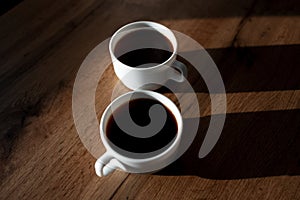 Close-up of two white ceramic cups of coffee with shadows on wooden table.