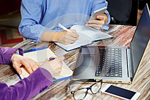 Close up of two women doing records in a notebook sitting at a laptop. Two pairs of women`s hands in an office.