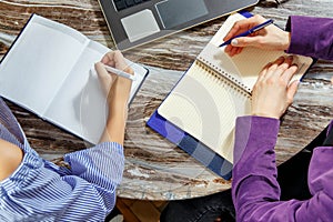 Close up of two women doing records in a notebook sitting at a laptop. Two pairs of women`s hands in an office. Laptop, glasses,