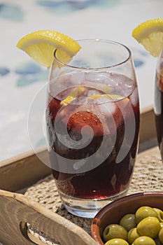 Close up of a typical spanish drinking glass of red wine with soda, ice cubes and slices of lemon next to a plate with olives on a