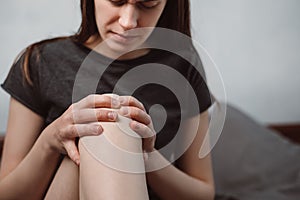 Close up of unhappy tired young woman sitting on bed at home massaging touching knee with hands, feeling pain.