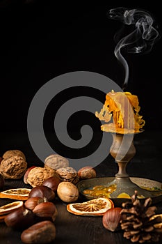 Close-up of unlit orange candle with smoke on dark wooden table with dried fruits and autumn leaves, black background