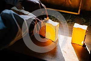 Close-up of unrecognizable apiarist cutting honeycomb using knife while standing in dark bee house.