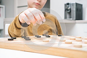 Close up unrecognizable cropped little boy hand with checker piece make step on wood board in kitchen. Strategy, logic