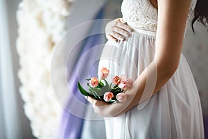 Close-up of unrecognizable pregnant woman with hands over tummy in white lace dresses with pink spring tulips