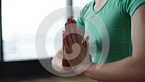 Close-up of unrecognizable spiritual woman holding joined palms at chest sitting in lotus pose on background of window.