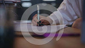 Close-up. An unrecognizable woman in a white blouse signs documents lying on her desk with a pen while sitting indoors.