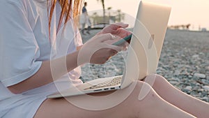 Close up of unrecognizable woman working with laptop and mobile phone on the beach by the sea during sunset. Female