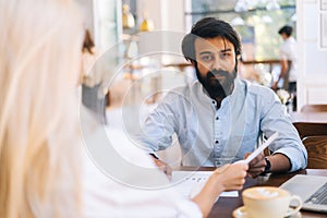 Close-up of unrecognizable young businesswoman manager passing papers documents to Indian colleague, sitting at table in