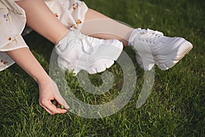 Close-up of unrecognizable young woman wearing white dress sitting on field with green grass at sunny summer day.