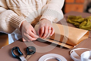 Close-up of unrecognizable young woman wrapping festive Christmas gift box in craft paper with presents for family and