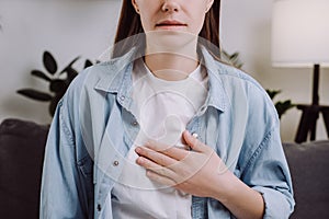 Close-up of upset unhealthy young female sitting on grey sofa at home having reflux acids and pain in chest. Gastroesophageal