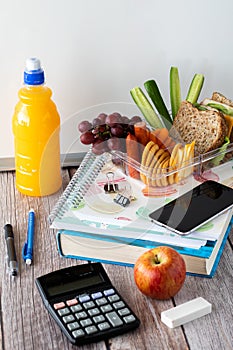 Close up of a variety of school supplies with a lunch kit on top and bottle of juice to the side, against a whiteboard.