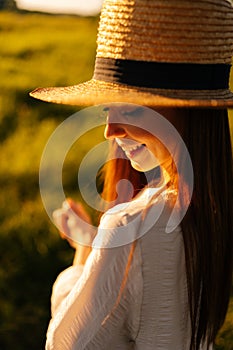 Close-up vertical face shot of smiling red-haired woman with closed eyes wearing straw hat and white dress standing