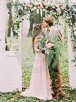 The close-up vertical photo of the groom in vintage suit kissing his bride with the big bouquet into the forehead under