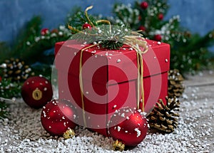 A close-up vertical photo of a red color gift box that stands on a wooden table among Christmas tree toys and coniferous branches
