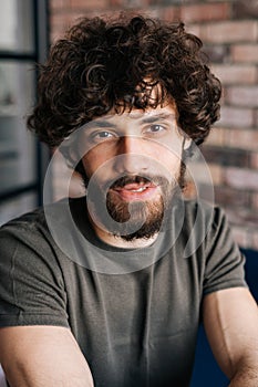 Close-up vertical portrait of friendly handsome young man looking at camera sitting on chair in cozy living room with