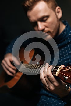Close-up vertical portrait of guitarist singer male playing acoustic guitar sitting on armchair in dark living room.