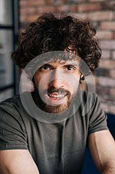 Close-up vertical portrait of handsome young man looking at camera sitting on chair in cozy living room with brick wall.