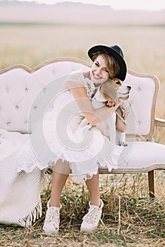 The close-up vertical portrait of the happy smiling bride hugging the fluffy dog while sitting on the cozy white sofa in