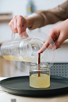 Close-up vertical shot of unrecognizable female artisan pouring melted wax into tranparent glass jar with wick at home.