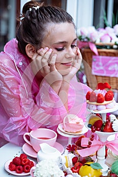 Close up vertical smiling dreaming teenage girl in pink raincoat joy and looking at sweet berry desserts in outdoor cafe