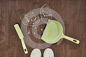 A close-up view from above of a broken transparent cup, brushes with a dustpan for cleaning and white sneakers. The