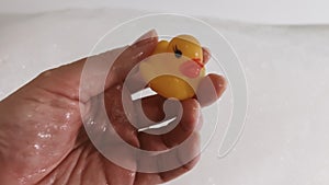Close-up view adult hand playing with yellow rubber duck. Toy duckling drowning in thick white soap bubble foam in