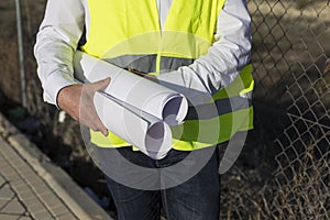 Close up view of Architect or Engineer man holding blueprints on the Construction Site. Wearing protection clothes. Crane on