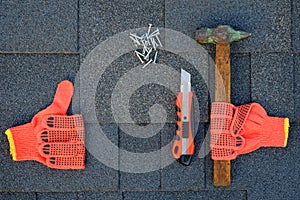 Close up view on asphalt shingles on a roof with hammer,nails and stationery knife. Use of gloves in construction.