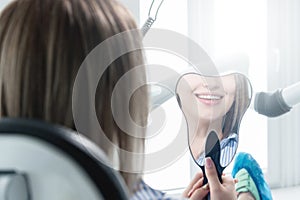 Close-up view from the back of a blonde girl sitting in a chair who looks in a tooth-shaped mirror. Reflection of a snow