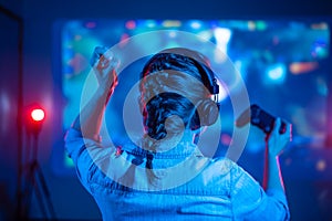 Close-up view from back of emotional gamer girl playing video game at home in front of big screen with joystick and headphone.