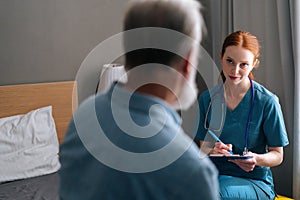 Close-up view from back of senior male patient talking with professional female doctor writing down diagnosis and