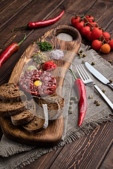 Close-up view on Beef Tartar with a raw egg yolk, rye bread and chilis on a wooden board and rough cloth with spoon and fork. Shot