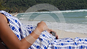 Close up view on the belly of a pregnant woman in dress sitting on beach bench.